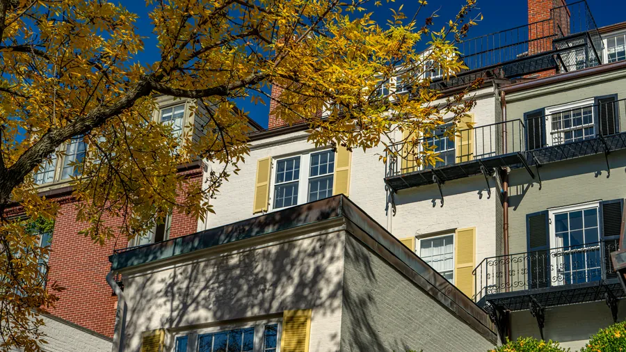 Federal-style brick and painted-brick homes with shutters and black iron balconies under a deep blue sky, framed by golden autumn leaves
