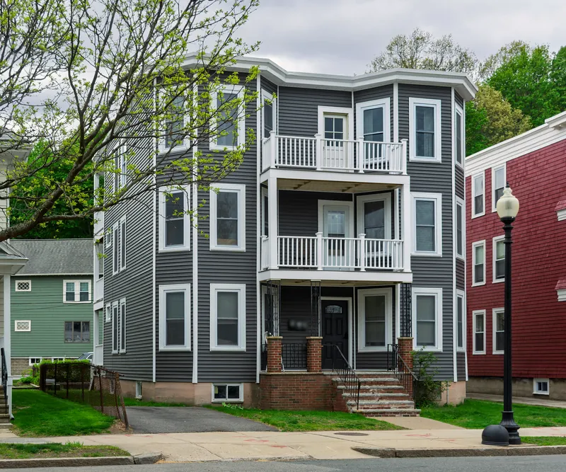 Renovated triple-decker with gray siding, white trim, stacked porches and a brick front stoop, framed by a young spring tree