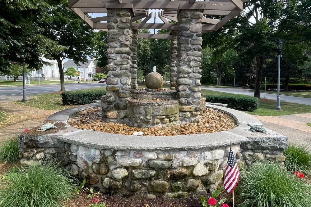 The stone C.S. Bird Fountain under a wooden pergola on the Walpole town common, surrounded by plantings